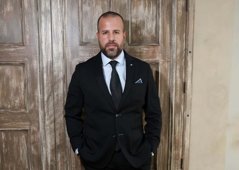 Man in a black suit with a tie and pocket square stands against weathered wooden doors, hands in pockets.